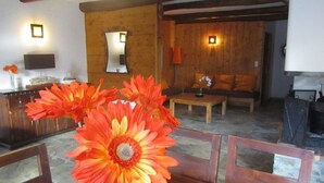 Ein gemütliches Wohnzimmer mit Holzboden, einer Couch, einem Couchtisch und einem Esstisch mit Stühlen. An den Wänden sind dekorative Lampen angebracht und auf dem Tisch steht eine Vase mit Blumen.