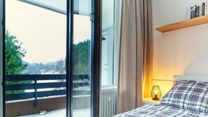A bedroom with a bed, a bedside table with a lamp, a window with curtains, and a view of a snowy landscape. - Apartment Winterberg - Stylish (Winterberg)