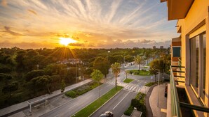 A sunset view from a balcony overlooking a road with trees and a building. - Destiny - Caparica apartment (Costa da Caparica)