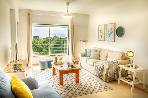 A well-lit living room with a large window, a sofa, a coffee table, and a side table with a lamp. - Destiny - Caparica apartment (Costa da Caparica)
