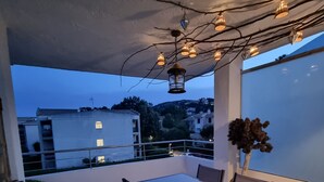 Un balcón con mesa y sillas, plantas en macetas y luces colgantes.