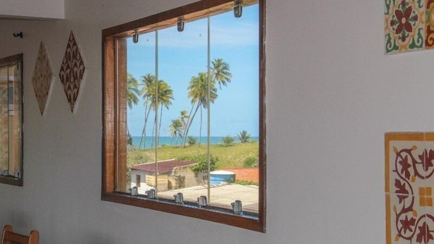 A room with a large window overlooking palm trees and the ocean. - Pousada Francês Praia (Marechal Deodoro)