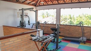 A gym with exercise equipment, a brick wall, and large windows with a view of palm trees. - Pousada Francês Praia (Marechal Deodoro)