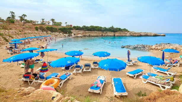 A beach with numerous blue umbrellas and sun loungers, a rocky shoreline, and clear blue water. - Kyklades Angie Resort (Paralimni)