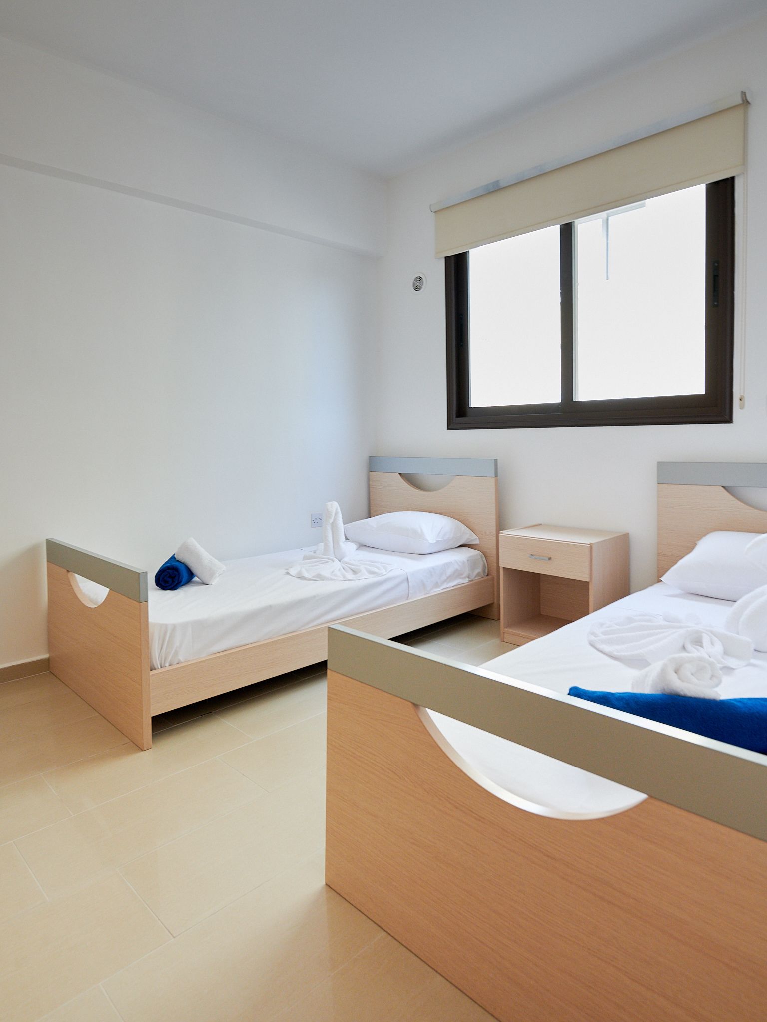 A hotel room with two single beds, a small bedside table, a window, and a light-colored tiled floor.