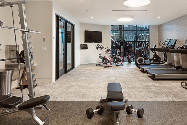 Un gimnasio con máquinas de cardio, bancos de pesas y un televisor en la pared.