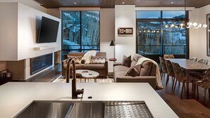 Une cuisine moderne dotée d’un grand îlot central, d’un coin repas avec une table et des chaises en bois, d’un canapé agrémenté de coussins à motifs et de grandes fenêtres offrant une vue sur une montagne enneigée.