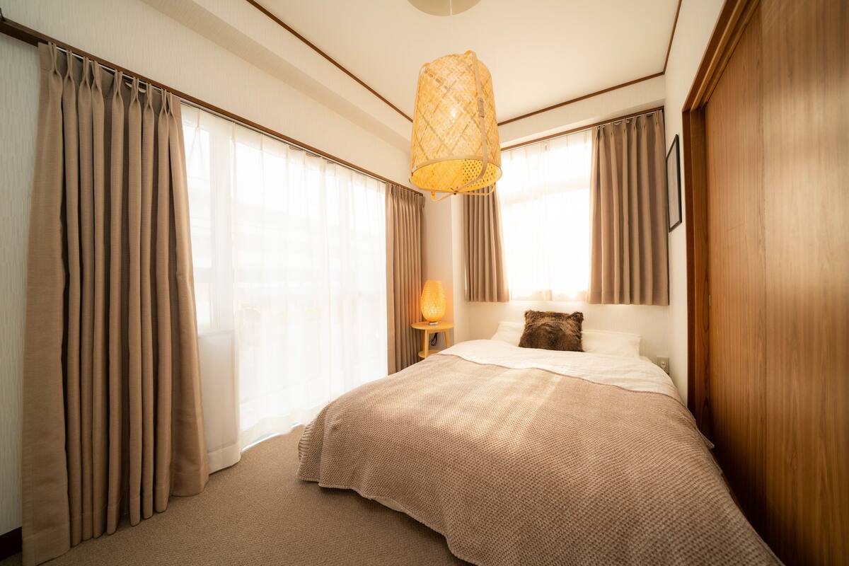 A hotel room with a large bed, beige curtains, a hanging light fixture, and a wooden wardrobe.