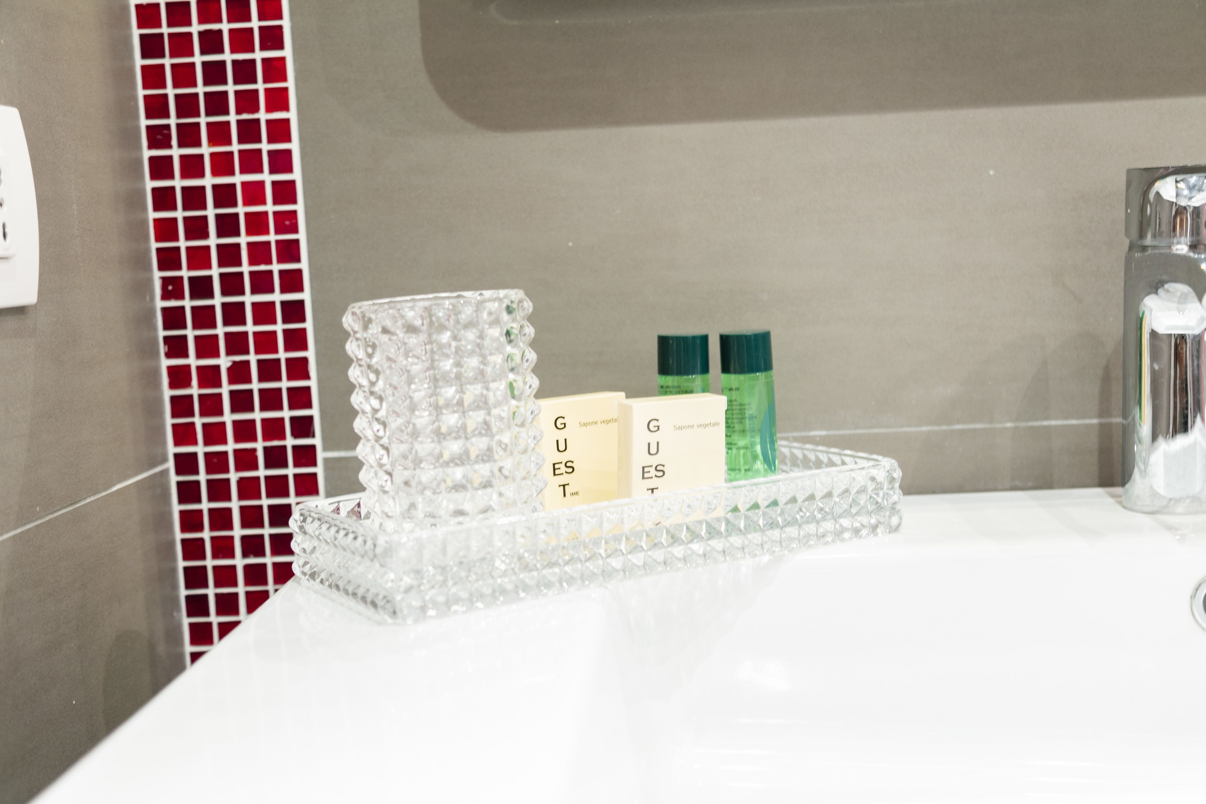 A bathroom with a white sink, a red mosaic tile backsplash, and a clear glass tray holding toiletries.