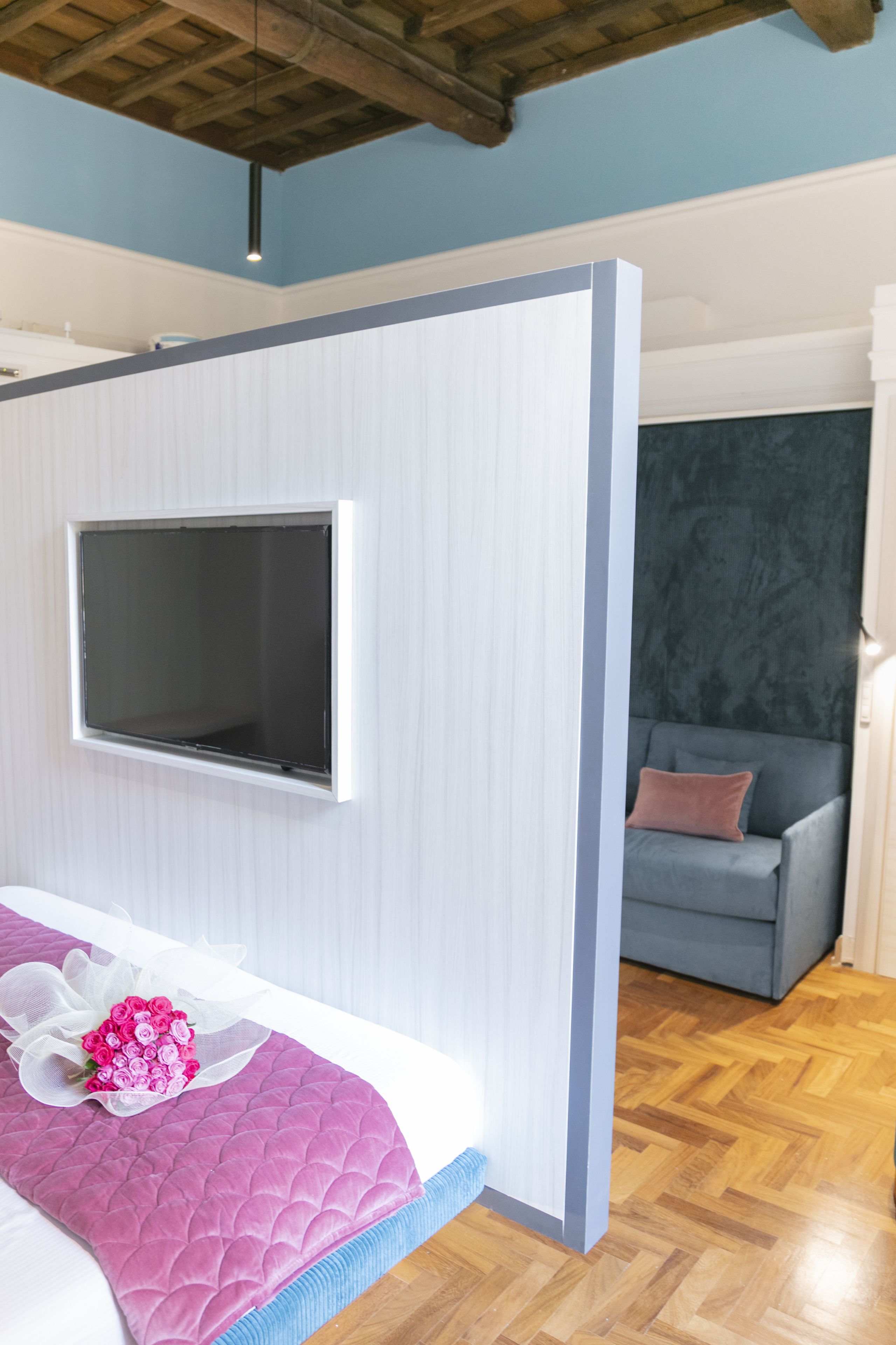 A bedroom with a bed, a TV mounted on the wall, a grey sofa, and a wooden floor.
