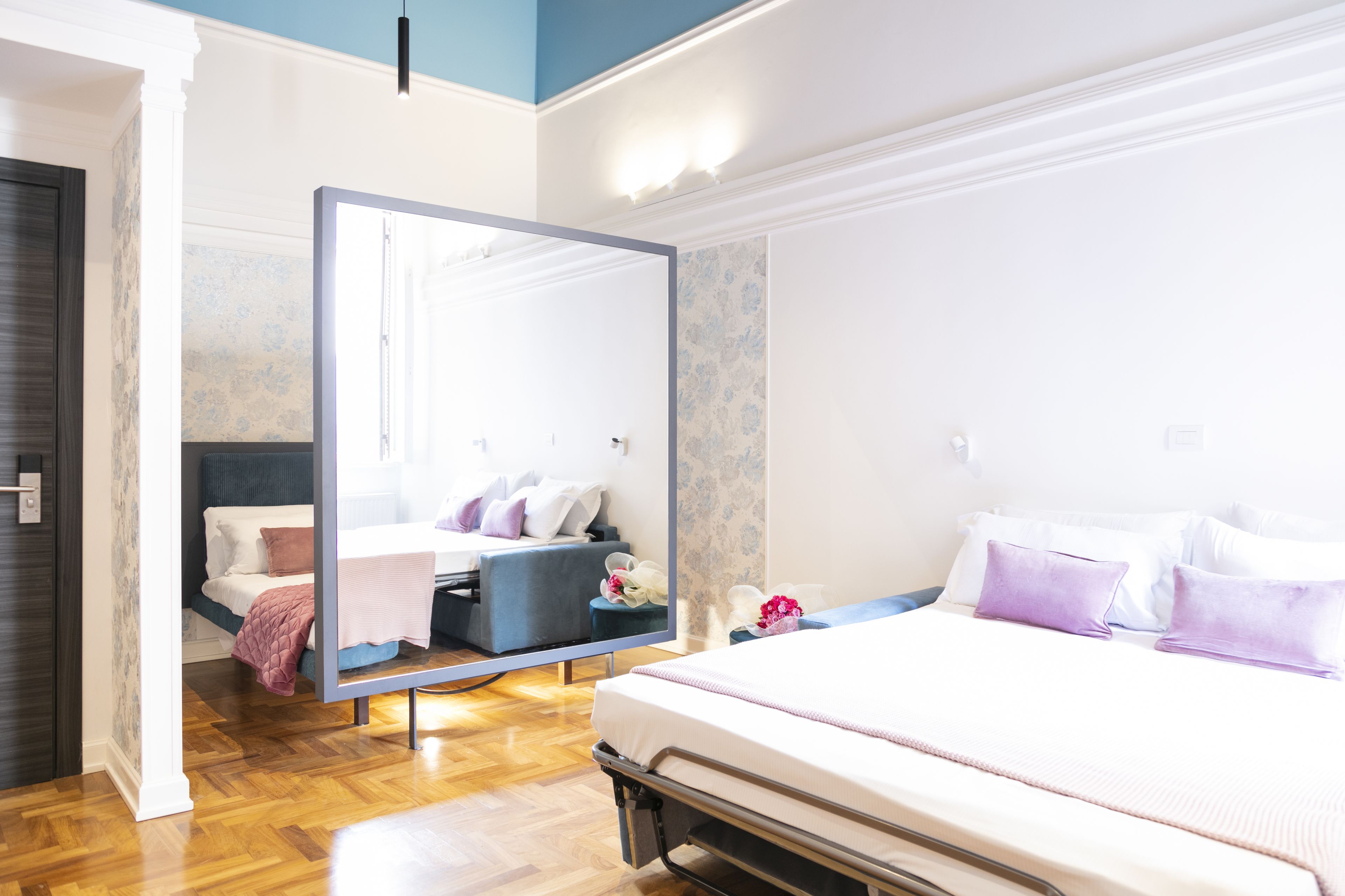 A modern hotel room with a large bed, a bedside table, a mirror, and a seating area with a sofa and chairs.