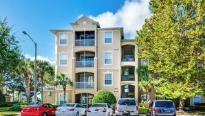 A multi-story apartment building with a beige exterior, balconies, and a parking lot with several cars. - EV_76604 - 3 Bed 2 Bath Condo with Access to Resort Amenities (Kissimmee)