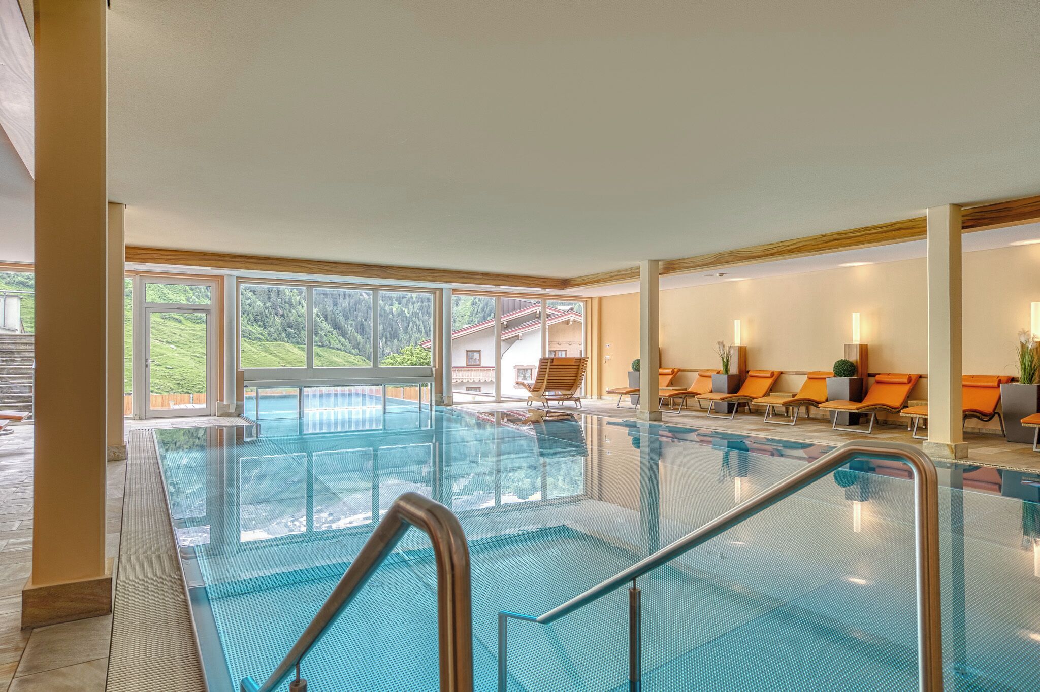 Une piscine intérieure avec des parois en verre transparent, un sol carrelé et des chaises longues orange.