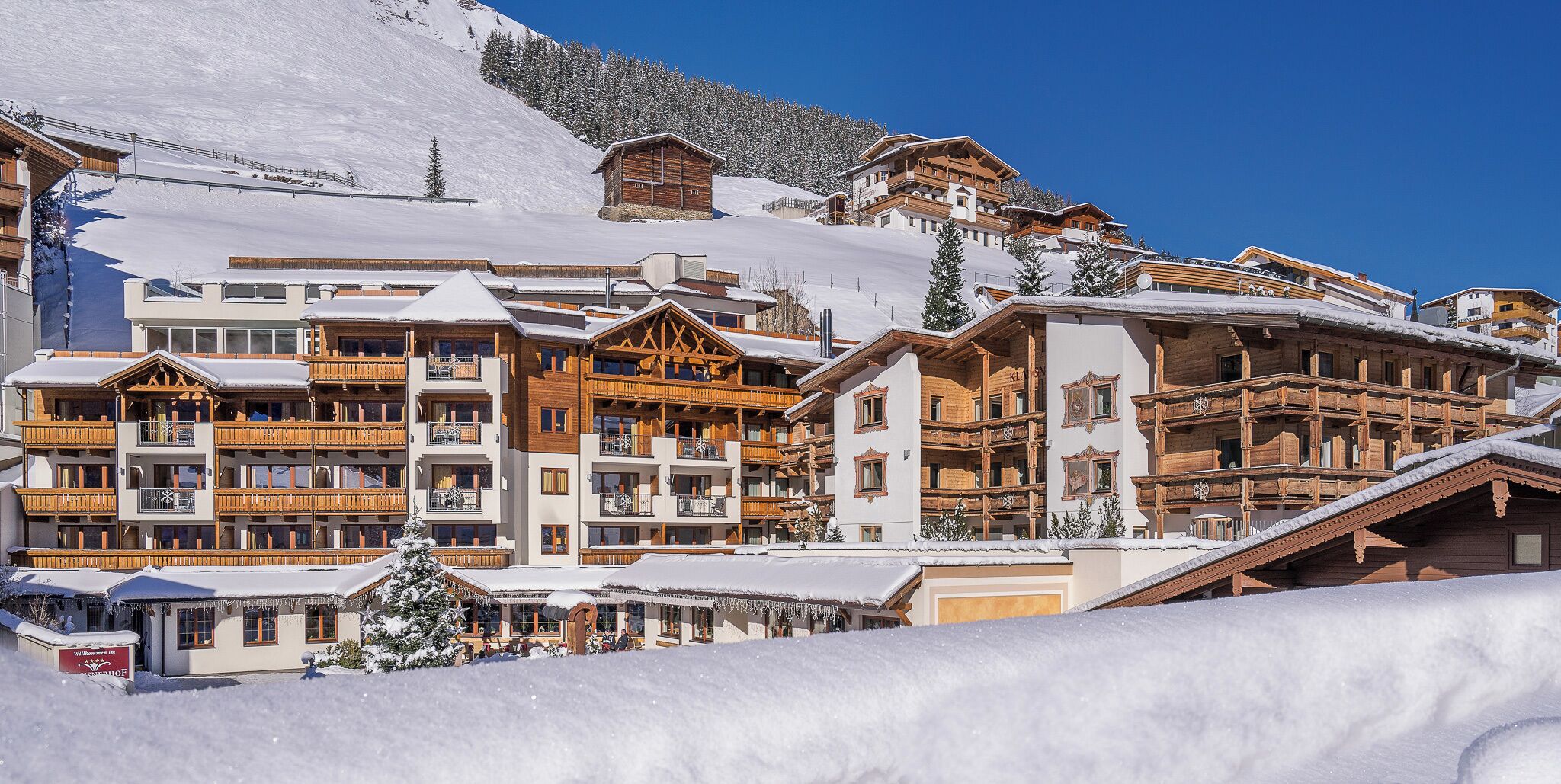 A snow-covered mountain resort with wooden buildings, balconies, and a ski slope in the background.