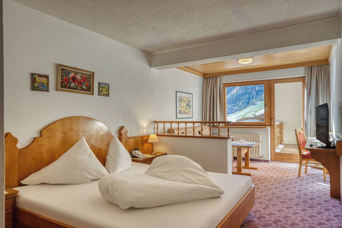 A hotel room with a large bed, a desk, a chair, a TV, and a window with a mountain view.