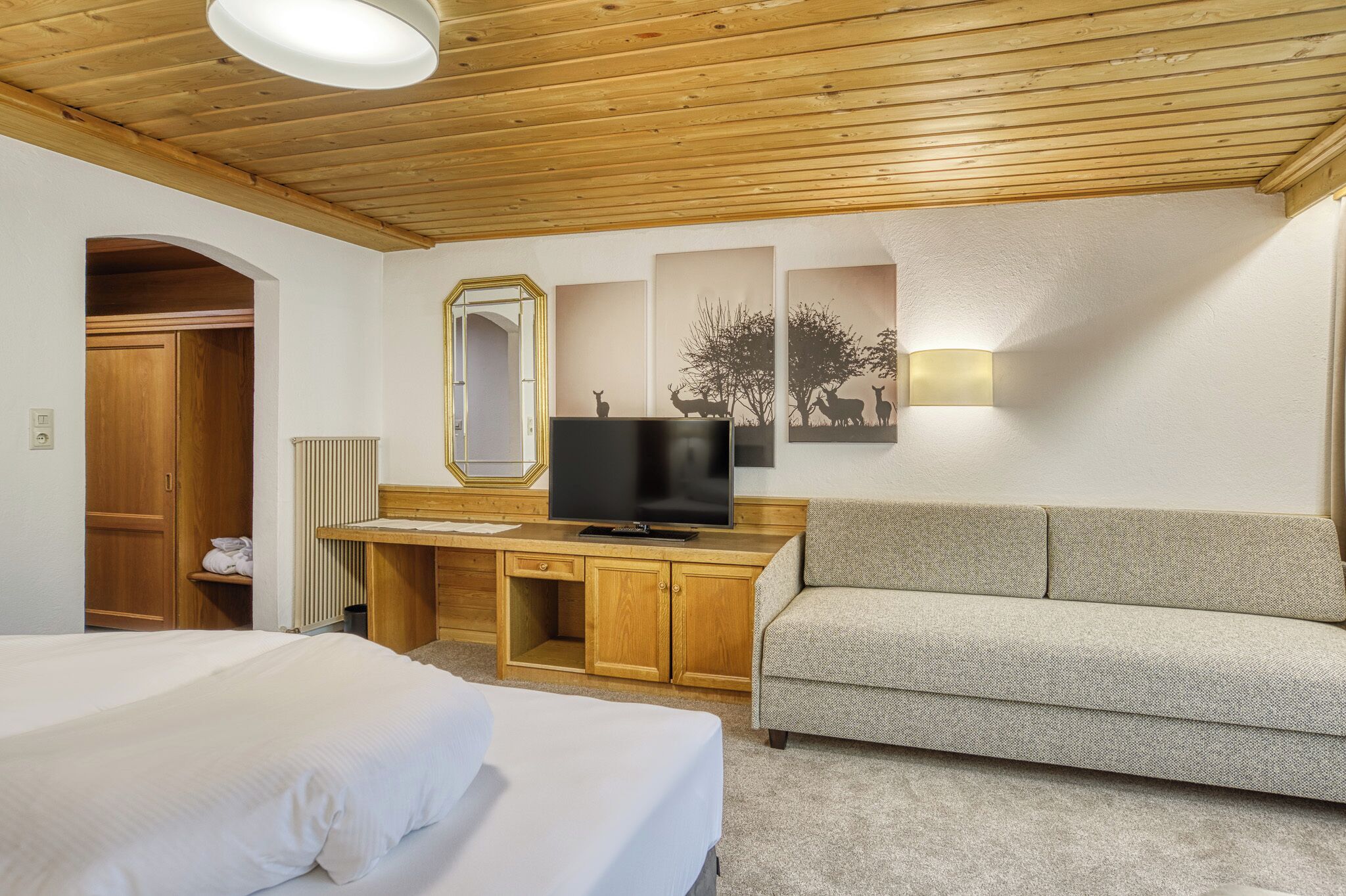 A hotel room with a wooden ceiling, a bed, a sofa, a desk with a TV, and artwork on the wall.