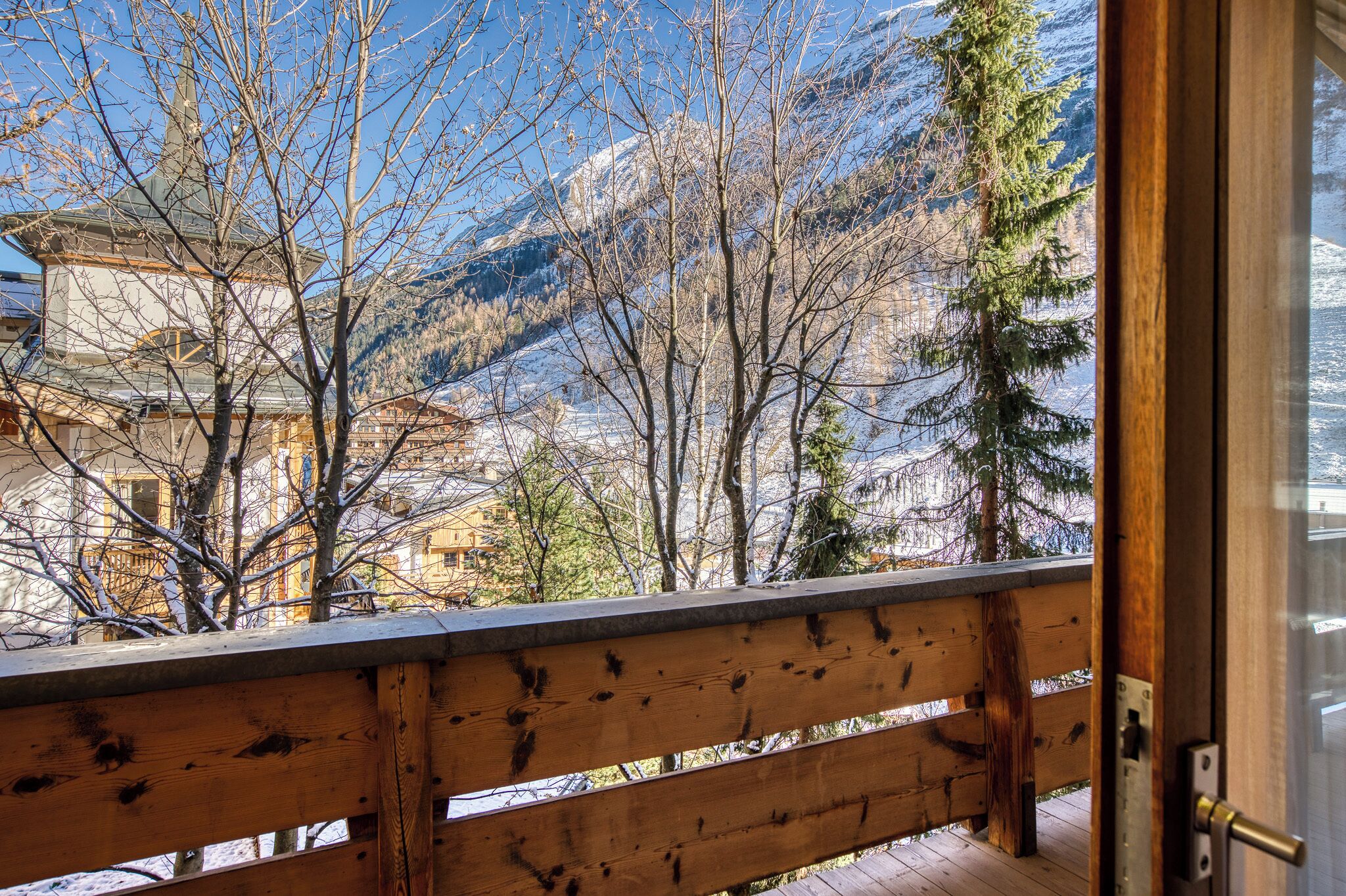 Un balcon en bois offrant une vue sur des montagnes enneigées et un bâtiment traditionnel doté d’un clocher.