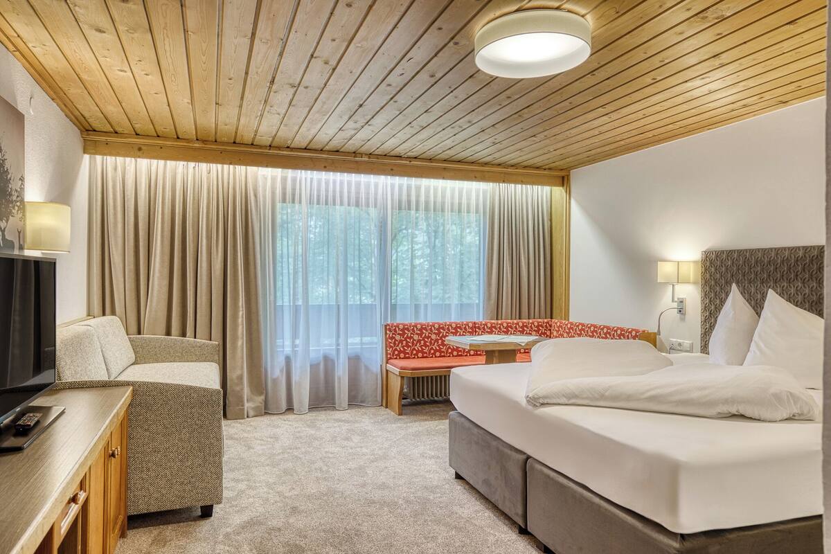 A hotel room with a wooden ceiling, a bed with white bedding, a desk with a television, and a window with curtains.