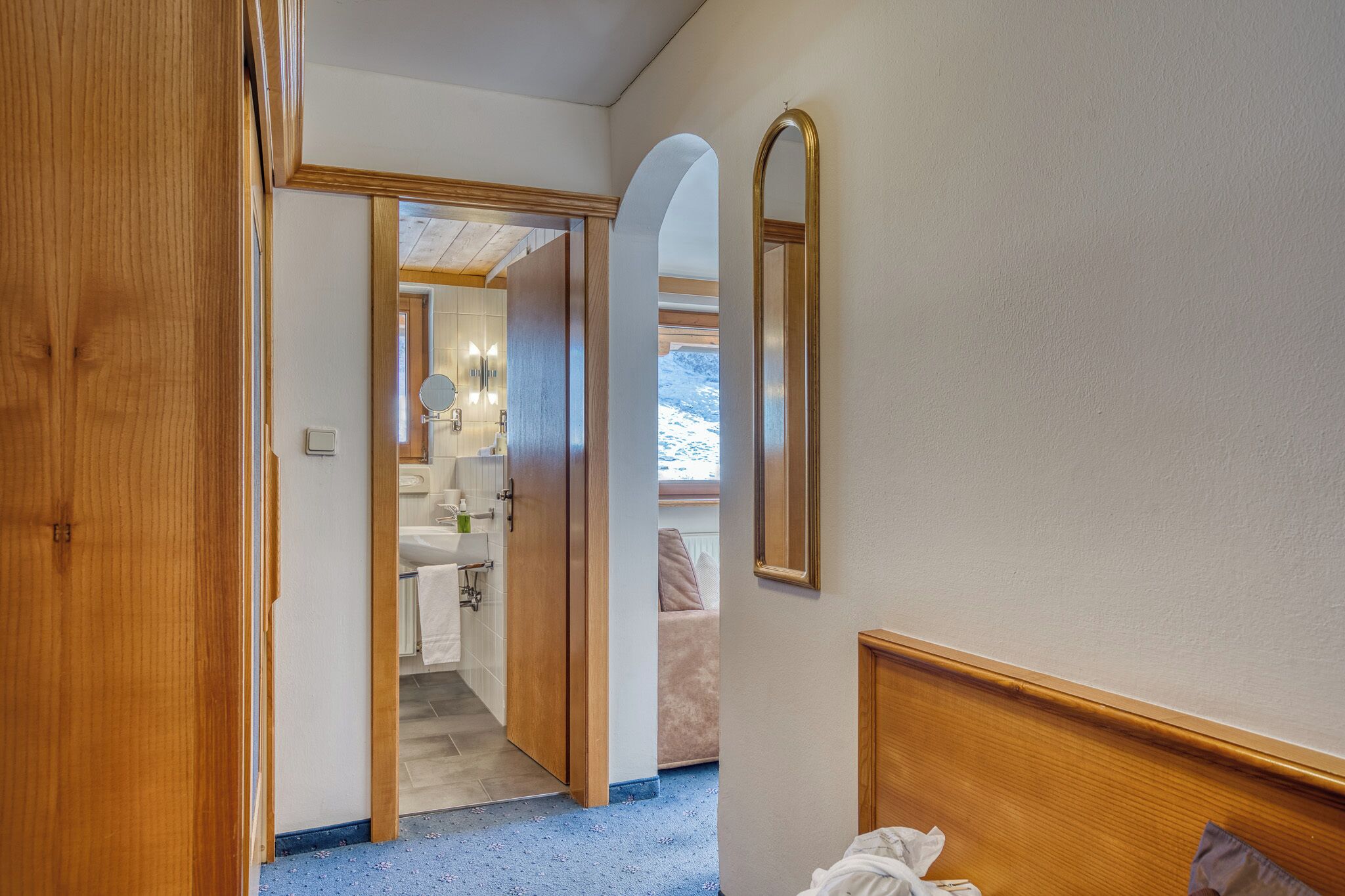 A hotel room with a wooden door, a mirror, a bed with a headboard, and a view of a bathroom with a sink and mirror.