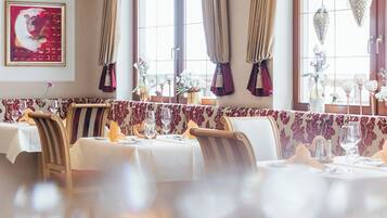 Restaurante com mesas postas para refeição, parede estampada e uma grande janela com cortinas.