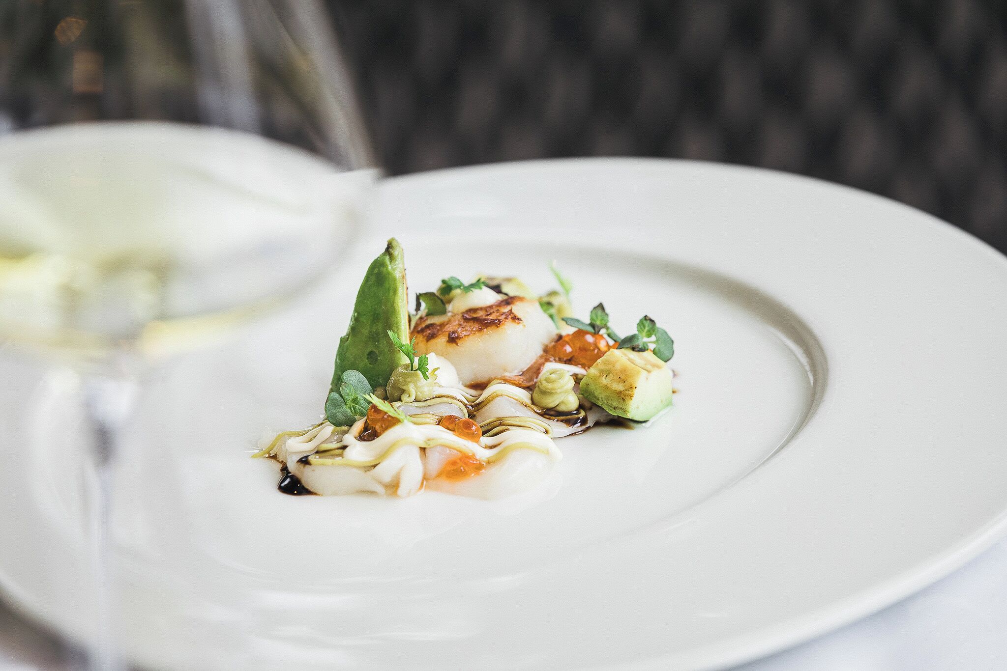 Plato blanco con un vieiras sellada, aguacate y guarniciones.