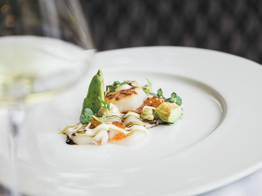 Ein weißer Teller mit gegrillter Jakobsmuschel, Avocado und Garnitur.