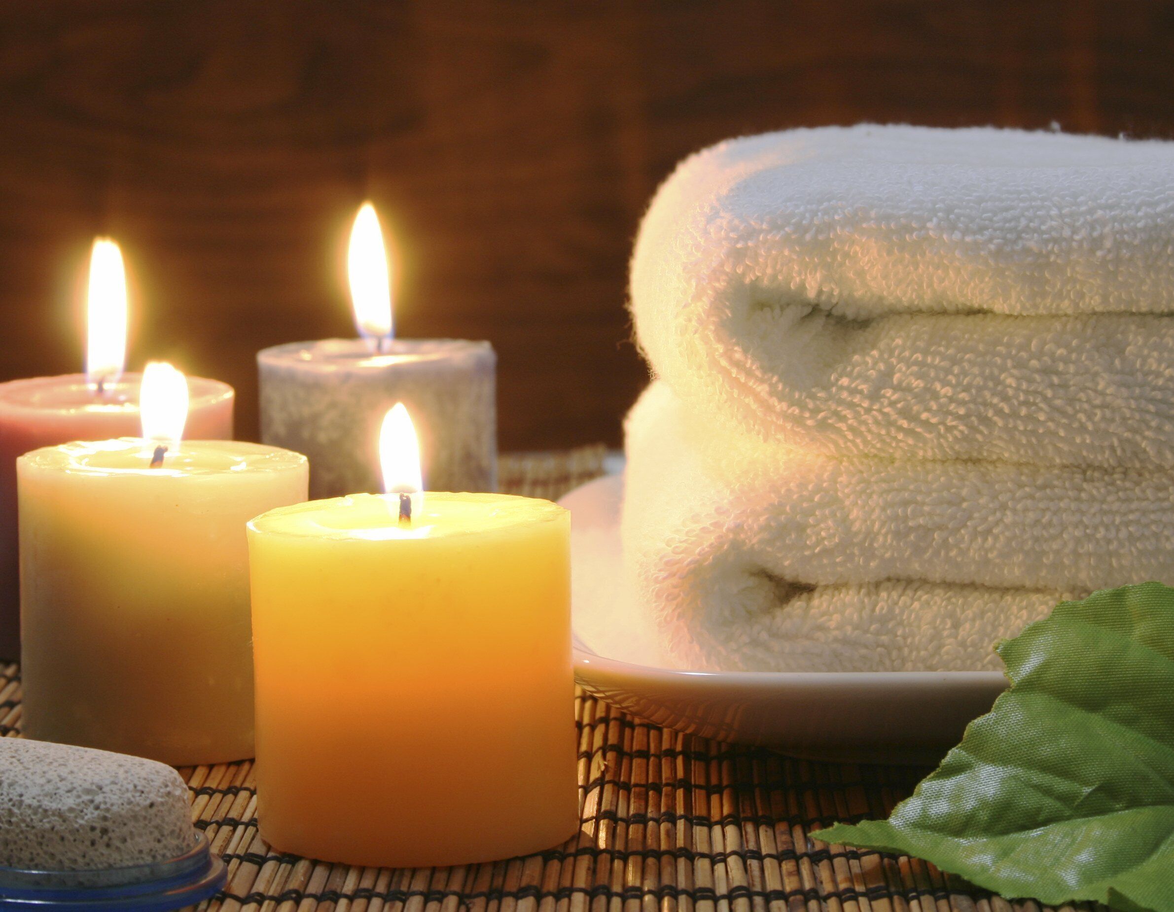 Three lit candles, a stack of white towels, and a green leaf on a bamboo mat.