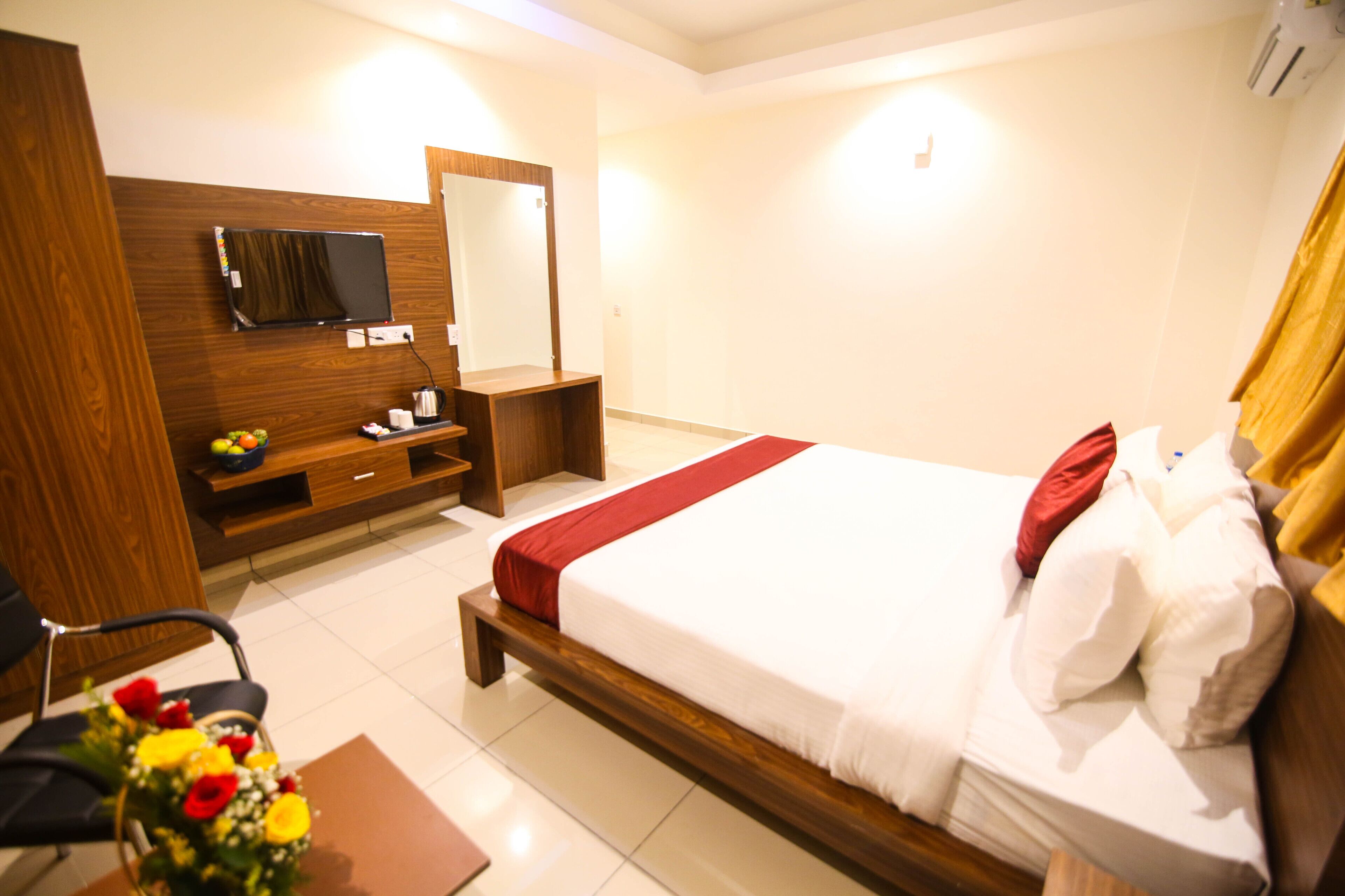A hotel room with a wooden bed, a desk with a computer, a TV mounted on the wall, and a small seating area with a chair and a table.