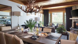A dining area with a wooden table set for four, surrounded by leather chairs. A chandelier with antlers hangs above, and a fireplace is visible in the background. - Outpost: Crystal Springs 306 - Ski-In/Ski-Out! (Teton Village)