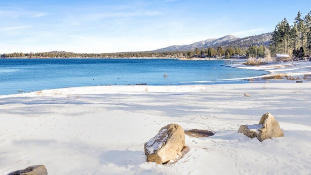 A snowy beach with clear blue water, a few rocks, and a forested mountain in the background. - Shore Acres Lodge #451 (Big Bear Lake)