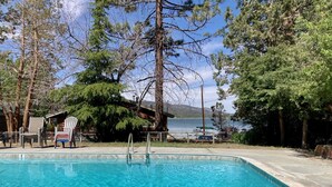 A clear swimming pool with a wooden deck, surrounded by trees and a view of a lake. - Shore Acres Lodge #451 (Big Bear Lake)