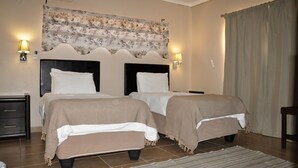 A bedroom with two beds, a headboard with a floral tapestry, bedside lamps, and a large window with curtains.