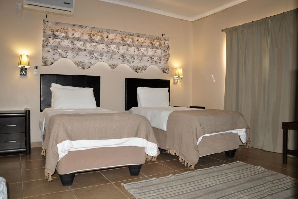 A bedroom with two beds, a headboard with a floral tapestry, bedside lamps, and a large window with curtains.