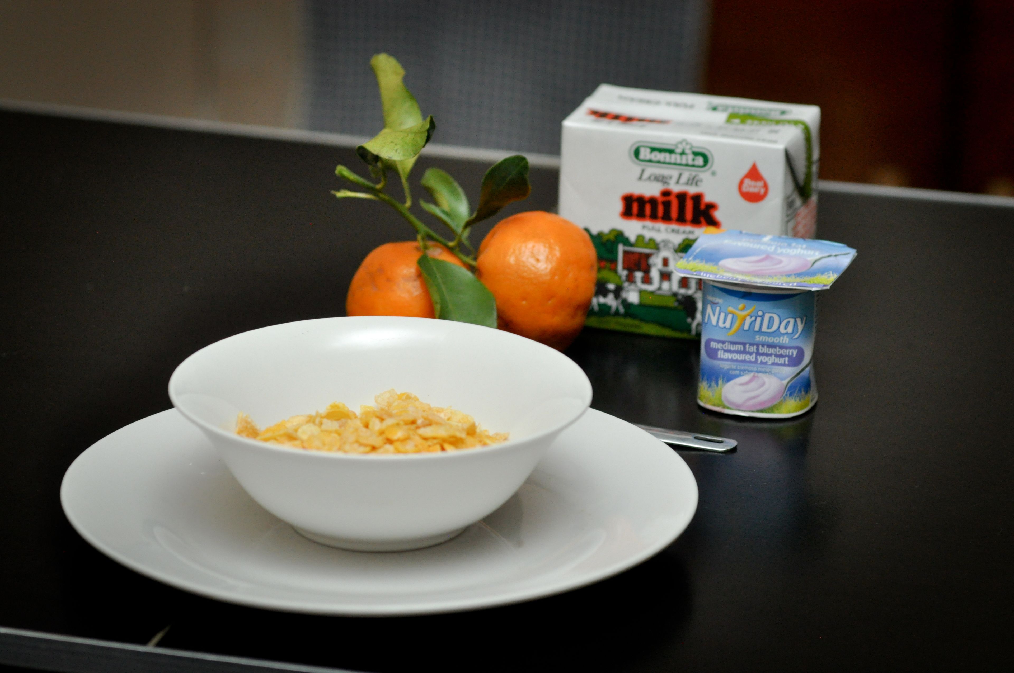 A bowl of scrambled eggs on a white plate, a carton of milk, a carton of NuYogi medium fat blueberry flavored yogurt, and a couple of oranges on a black table.