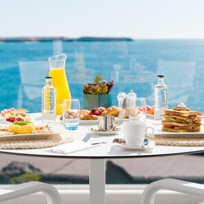 Une table de petit-déjeuner, dressée avec divers aliments et boissons, avec en toile de fond une vue sur la mer.