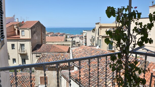 Une vue sur des toits et des bâtiments recouverts de tuiles en terre cuite, avec un ciel bleu dégagé et la mer en arrière-plan.