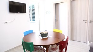 Un coin repas avec une table ronde, des chaises colorées, un panier de pain et un téléviseur fixé au mur.