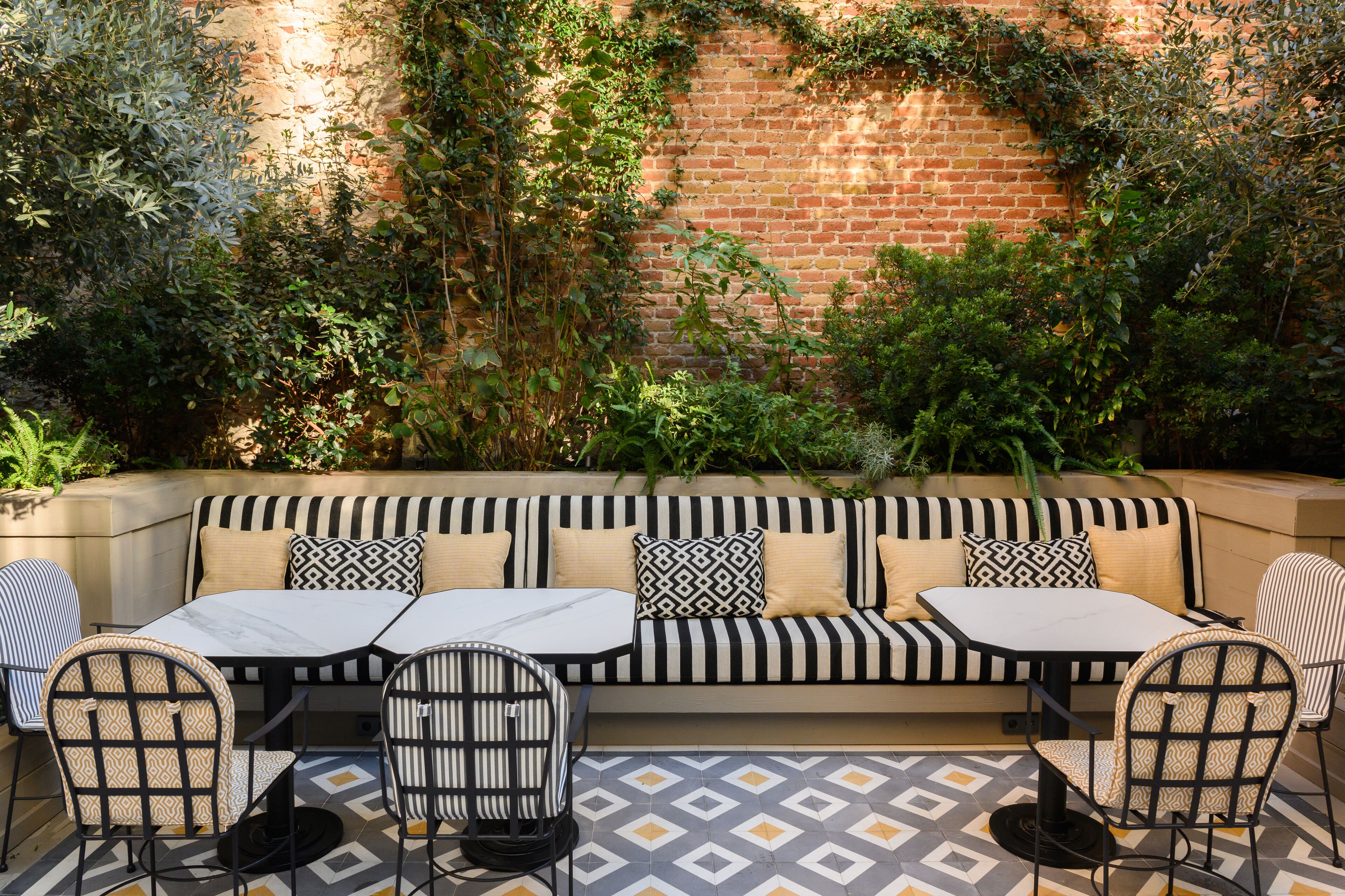 A brick wall with climbing plants, a striped sofa with patterned cushions, and a tiled patio with two tables and chairs.