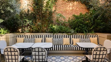 A brick wall with climbing plants, a striped sofa with patterned cushions, and a tiled patio with two tables and chairs.