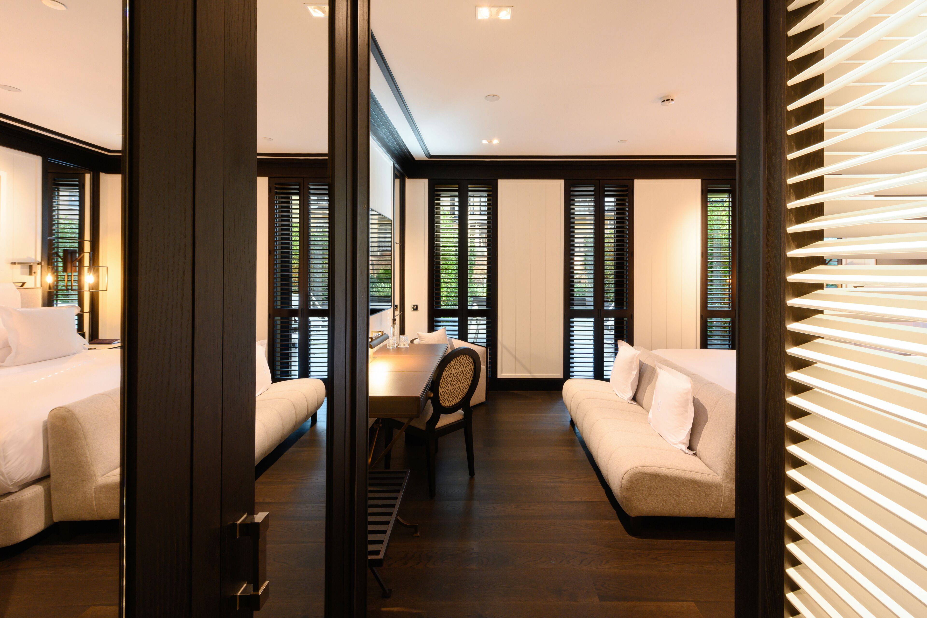 A modern hotel room with a dining area, two beds, and a large window with blinds.