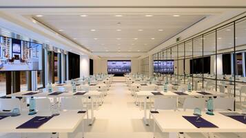 A modern conference room with white tables, chairs, and a large screen at the front.