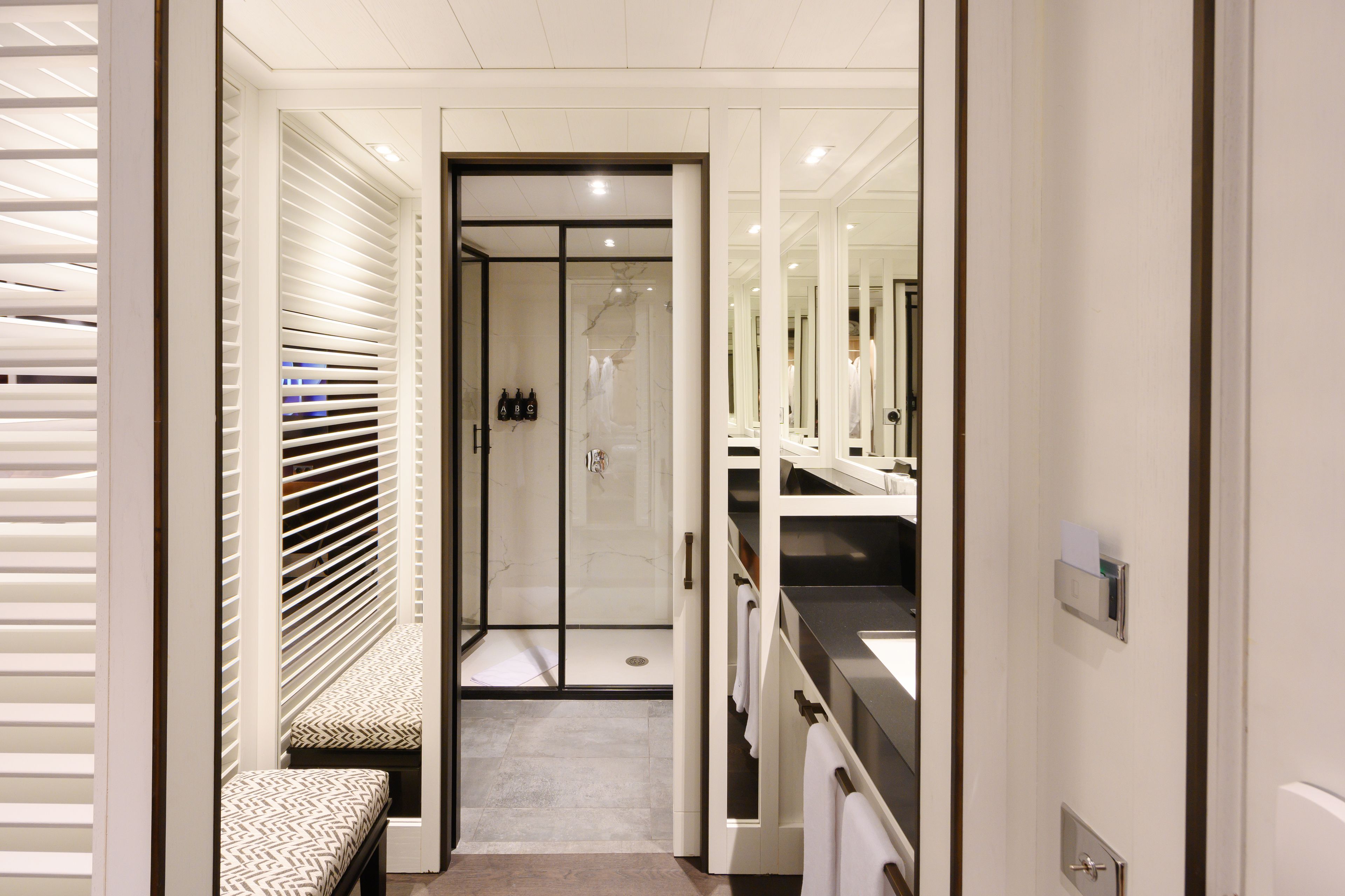 A narrow hallway with a bench, a glass door leading to a bathroom, and a vanity with a mirror and towel rack.