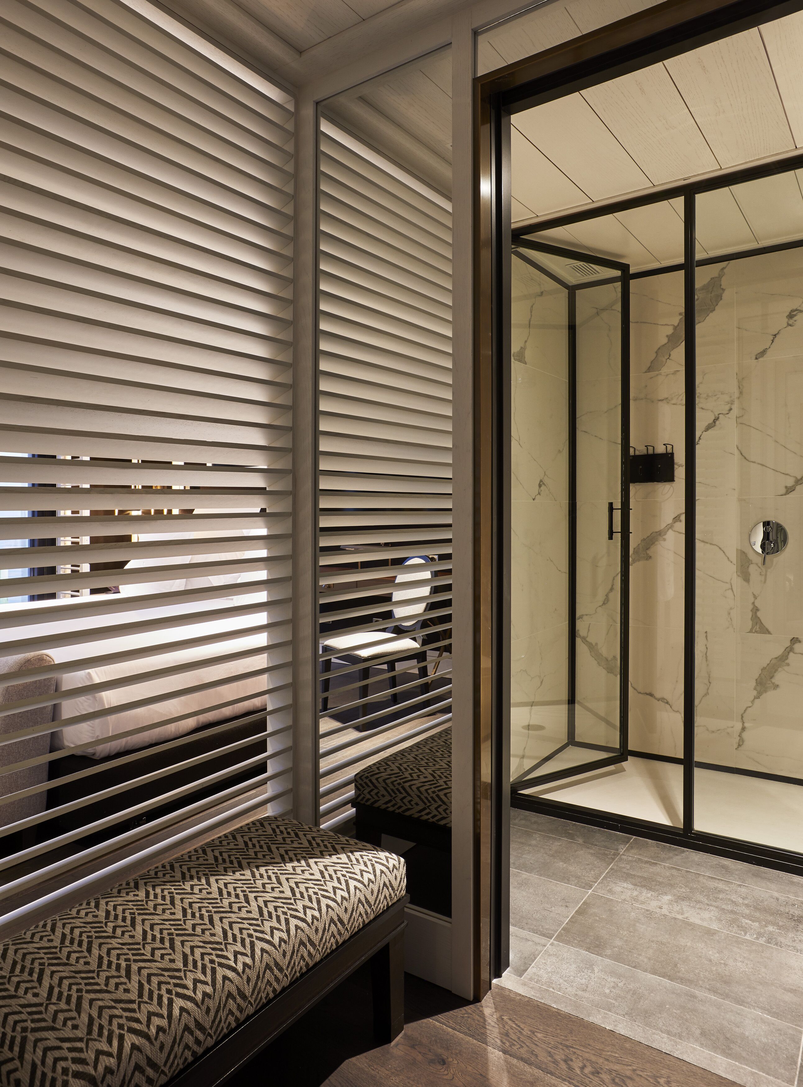 Un baño moderno con cabina de ducha de vidrio, un banco con asiento estampado y una pared con persianas horizontales.