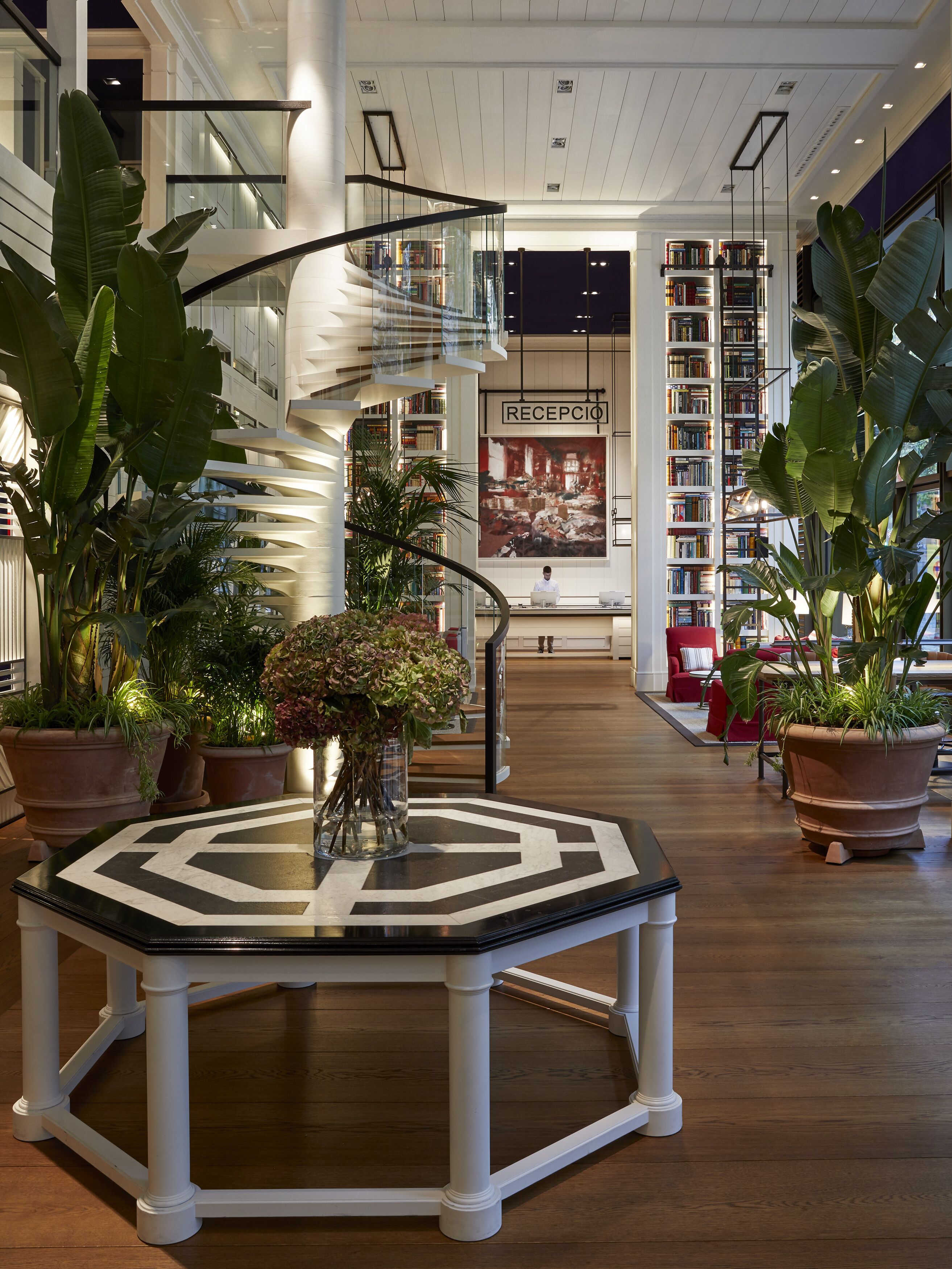 Um hall amplo com uma mesa octogonal central, rodeado por plantas em vasos, e uma escadaria em espiral que leva aos andares superiores.