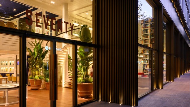 Un edificio moderno con fachada de vidrio, grandes macetas y un interior visible con asientos y mesas.