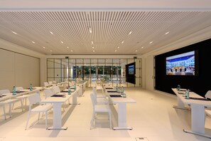 A modern conference room with white tables and chairs, a large screen, and a view through glass walls.