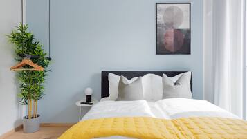 A neatly made bed with a yellow blanket, a potted plant, and a wall-mounted artwork.