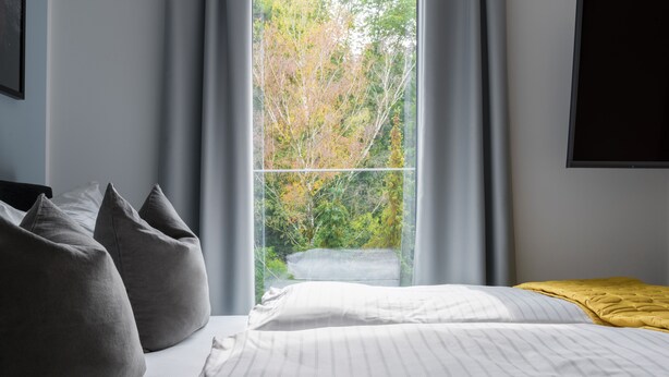 Un dormitorio con cama, almohadas, una ventana con cortinas y vistas a árboles.