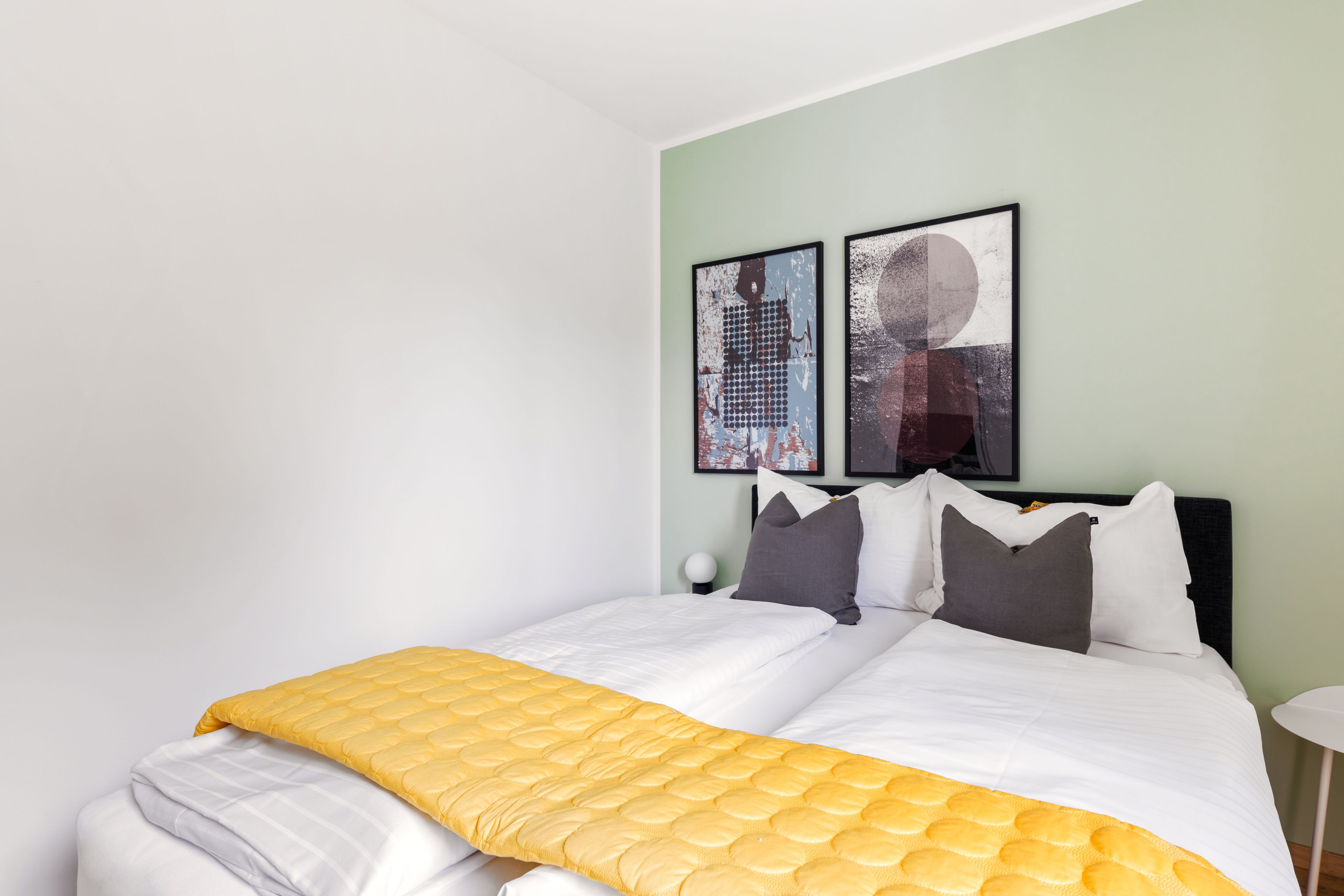 A neatly made double bed with a yellow quilt, white linens, and gray pillows. There are two framed artworks on the wall above the bed.