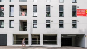 Un edificio moderno con múltiples ventanas y balcones, que presenta una entrada de vidrio grande y un letrero rojo a la derecha.